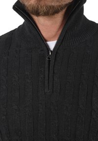 Schwarzer Strickpullover mit hohem Kragen und Reißverschluss bis zur Hälfte. Verfügt über ein Zopfmuster und eine strukturierte Stoffoberfläche. Lässige Passform.