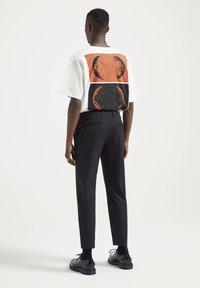 Homme portant un t-shirt blanc avec des motifs circulaires orange et noirs dans le dos, un pantalon noir et des chaussures habillées noires, debout de dos.