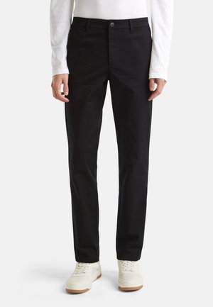 Chinos de algodón negro con un diseño de pierna recta, cinturilla con trabillas y un cierre frontal de botón único. Llevados con zapatillas blancas.