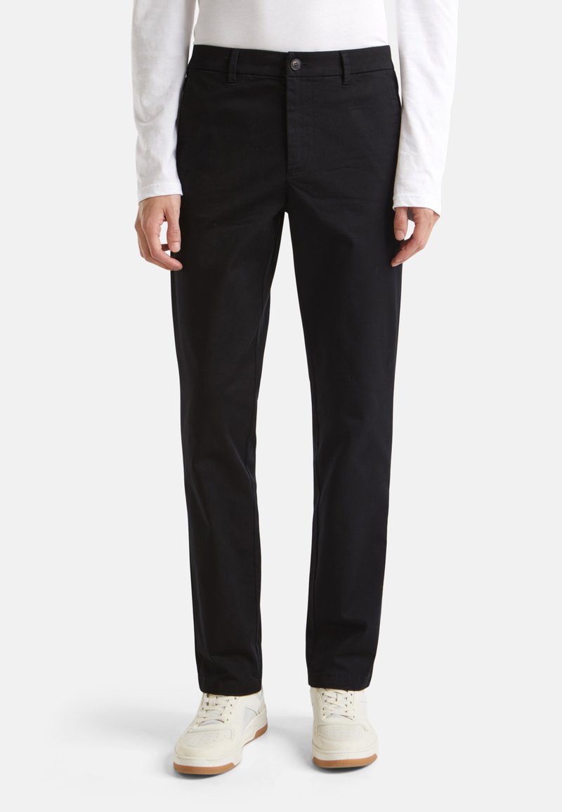 Chinos noirs en coton avec une coupe droite, une ceinture avec passants, et une fermeture à un bouton à l'avant. Portés avec des baskets blanches.