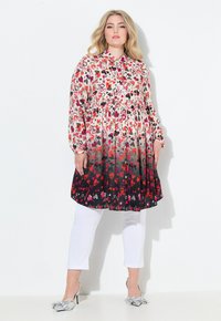 Abito a campana con maniche lunghe, colletto e motivi a cuori in rosso e nero, che si sfumano in una tonalità più chiara nella parte superiore; abbinato a pantaloni bianchi.