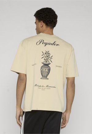 Ung mann med krøllete hår sett bakfra, iført beige T-skjorte med svart illustrasjon av en blomstervase og tekstene "Pegador" og "Moments to Memories."