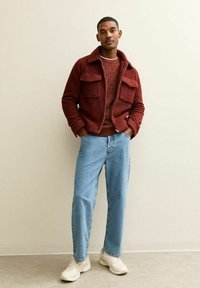 Bordeaux fleece jack met twee borstzakken, gestreepte bruine trui, lichtblauwe jeans en beige sneakers, staand tegen een lichte muur.