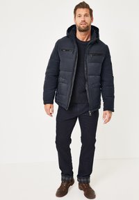 Marineblaue Steppjacke mit Kapuze, Reißverschlussfront und Brusttaschen; kombiniert mit dunklen Hosen und braunen Schuhen, die mit einem karierten Manschettendetail versehen sind.