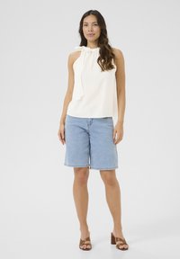 Haut sans manches crème léger avec un col froncé et un détail à nouer, associé à un short en denim et des sandales à talons marron.
