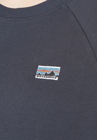 Sudadera de algodón gris oscuro que presenta un parche rectangular con el logo de Patagonia, con gráficos de montañas y acentos en azul y rojo.