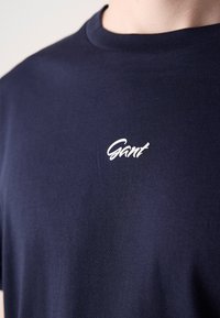 Tricou bleumarin din bumbac, cu gât rotund și un logo alb "Gant" brodat în zona pieptului. Textură moale.