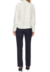 Blouse en soie blanche avec un col montant et des poignets boutonnés, associée à un pantalon taillé bleu marine foncé et des escarpins noirs. Coupe lisse et structurée.