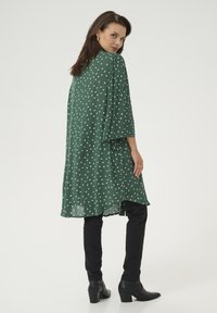 Robe verte à pois blancs, coupe ample, manches trois-quarts, ourlet asymétrique. Associée à un pantalon slim noir et des bottines.