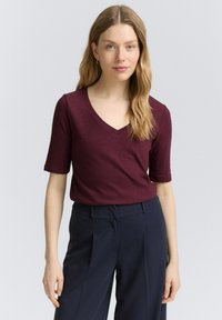 Bordeaux V-hals t-shirt, korte mouwen, zachte stof, gecombineerd met marineblauwe broek, gepresenteerd tegen een lichtgrijze achtergrond.