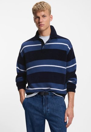 Knitted pullover with a dark blue and light blue striped pattern, featuring a collared neckline and long sleeves. Paired with blue jeans.