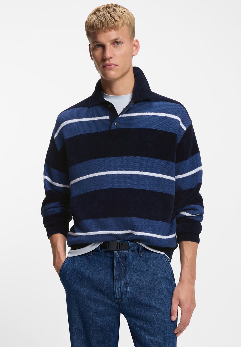 Knitted pullover with a dark blue and light blue striped pattern, featuring a collared neckline and long sleeves. Paired with blue jeans.