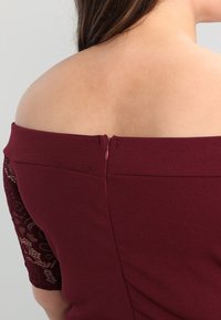 Burgunderfarbenes schulterfreies Kleid mit kurzen Spitzenärmeln und einem Reißverschluss am Rücken. Glatter Stoff mit figurbetontem Design.