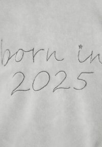 Világosszürke szövet, amelyen hímzett szöveg található: "2025-ben született" ívelt, dőlt betűtípussal, a "ben" szó mellett egy kis csillag díszítéssel.