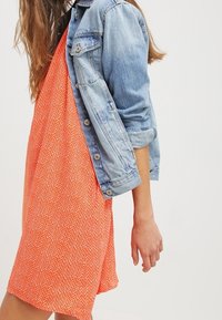 Jeansjacke mit hochgekrempelten Ärmeln über einem leuchtend orangefarbenen, geometrisch gemusterten Kleid. Das Kleid ist ärmellos und hat eine lockere Passform.