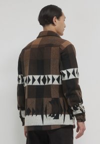 Veste à carreaux brune et noire avec des accents géométriques blancs, poignets à boutons et col. Matériau en laine texturée, design mi-long.