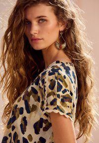 Ovratnik z leopardjim vzorcem v kremasti, zeleni in modri barvi, z valovitimi rokavi. Zlati uhani z rdečimi poudarki in zapletenimi vzorci, ob valovitih laseh.