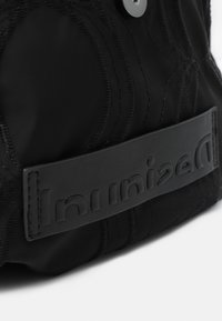 Primer plano de tela negra con patrones cosidos y una etiqueta de cuero negro grabada en relieve con el texto "Desigual" invertido.