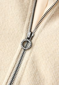 Beżowa dzianinowa kurtka zapinana na zamek, z teksturowaną powierzchnią, ozdobiona srebrnym zamkiem błyskawicznym i okrągłym uchwytem z logo.