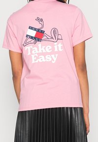 Růžové bavlněné tričko s kresleným designem na zádech, nápisem "Take it Easy" a logem značky, kulatým výstřihem a krátkými rukávy.