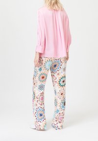 Femme portant un blouse rose ample et un pantalon blanc à jambes larges avec des motifs circulaires tie-dye colorés, se tenant de dos.