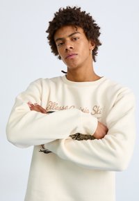 Sudadera color crema con mangas largas, que presenta un estampado gráfico y texto en marrón. Tejido suave con un corte holgado y diseño de cuello redondo.