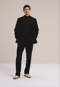 Schwarzer Anzug mit einem eng anliegenden schwarzen Rollkragenpullover, der ein maßgeschneidertes Design aufweist. Dazu graue und weiße Sneaker, vor neutralem Hintergrund.