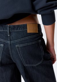 Jean en denim foncé avec une étiquette en cuir marron à l'arrière, doté de coutures blanches et de poches classiques. Coupe légèrement ample, design décontracté.