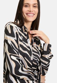 Schwarze Bluse mit abstrakten Wellenmustern in Creme, Beige und Grau; verfügt über einen Kragen und Knopfmanschetten, aus glattem Stoff gefertigt.