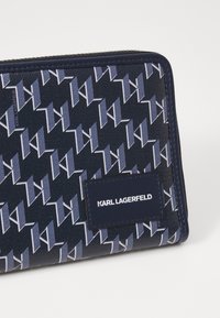 Jūrų laivyno piniginė su geometriniu raštu mėlynų atspalvių. Pagaminta iš odos, su stačiakampio formos logotipo etikete "KARL LAGERFELD". Uždarymas užtrauktuku.