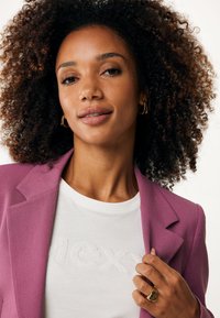 Blazer lilas sur un t-shirt blanc texturé avec "MEXY" brodé. Boucles d'oreilles en or et une bague en or accentuent le look. Cheveux doux et bouclés.