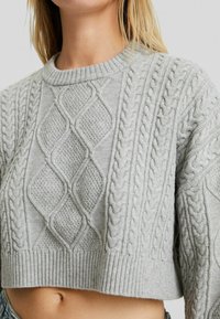Kort lichtgrijs gebreid sweater met diamant- en vlechtpatronen, gedragen door een persoon met blond haar en een blote buik.