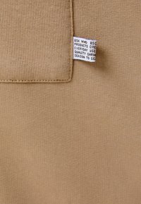 Beige Stoff mit einer glatten, weichen Textur zeigt ein sichtbares Pflegeetikett. Das Etikett enthält Pflegehinweise für Textilien und Markeninformationen.