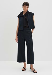 Haut sans manches noir avec une capuche attachée au cou, associé à un pantalon large noir et des sandales à talons hauts noires, sur fond blanc.