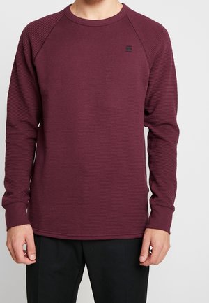 Mężczyzna ubrany w bordową długorękawową bluzę o fakturowanej powierzchni z czarnymi spodniami na jednolitym tle.