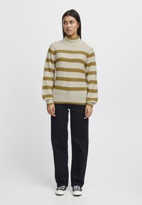 Gestreifter Pullover aus Strick in Beige und Olivgrün, mit hohem Kragen und langen Ärmeln, kombiniert mit schwarzen Hosen und lässigen Schuhen.