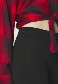 Leggings noirs avec une large ceinture texturée, assortis à une chemise à carreaux rouge et noir, boutonnée et nouée à la taille.