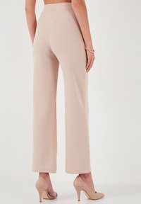 Beige, bő szárú nadrág, texturált anyagból, magas derékkal. Meztelen színű magassarkúval párosítva. A ruha hátsó nézete.