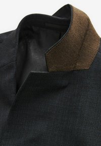 Dunkelblazer mit strukturiertem Finish, ausgestattet mit einem braunen Filzkragen und subtilen Nähten. Das Innere hat ein gemustertes Futter.