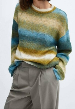 Vrouw in een losse, gestreepte gebreide trui in blauw, groen, beige en wit met lichtgrijze high-waisted pantalon, handen in de zakken.