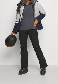 Skioutfitet består av en grå puffy-jacka, en mörkgrå fleece-tröja, svarta byxor och svarta skidskor. En svart hjälm hålls i ena handen.