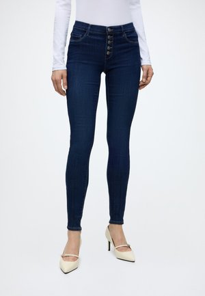 Person iført mørkeblå højttaljede skinny jeans med flere knapper og cremefarvede spidse stiletter med ankelstropper.