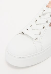 Vit sneaker med en slät läderöverdel, med vita snören, silverfärgade öglor och en metallisk roséguldig detalj på sidan.