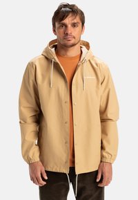 Beige waterdichte jas met een capuchon, elastische boorden en drukknoopsluiting. Witte logo op de borst. Draagt over een oranje shirt.