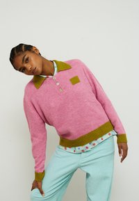 Vaaleanpunainen neulottu pusero, jossa on vihreä kaulus ja helma, kolme nappia ja neliskanttinen merkki. Yhdistetty vaaleansinisiin resorihousuihin.