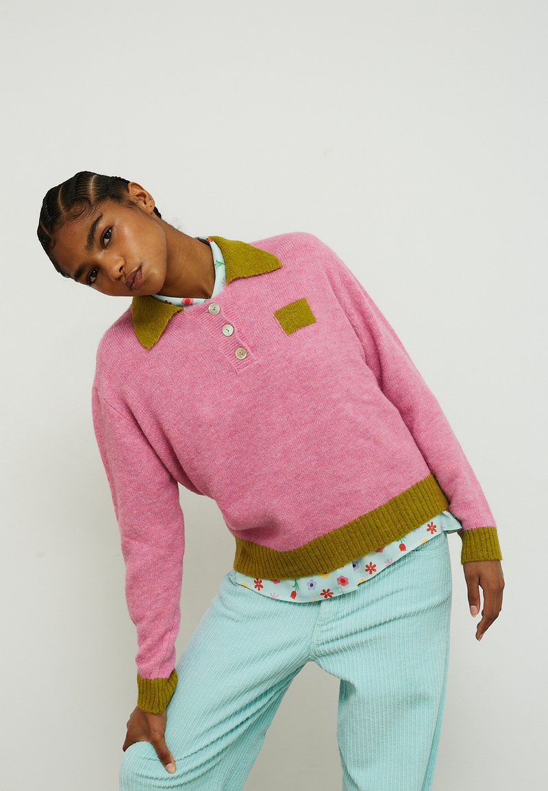 Vaaleanpunainen neulottu pusero, jossa on vihreä kaulus ja helma, kolme nappia ja neliskanttinen merkki. Yhdistetty vaaleansinisiin resorihousuihin.