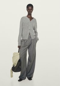 Donna che indossa un cardigan grigio con bottoni e pantaloni larghi grigio a righe, tiene in mano una borsa marrone con un maglione beige appoggiato sopra.