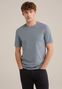 Camiseta de algodón de color azul claro con mangas cortas, cuello redondo y textura suave. El modelo posa con las manos en los bolsillos, vistiendo pantalones oscuros.