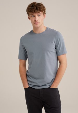 T-shirt en coton bleu clair à manches courtes, décolleté rond et texture lisse. Le modèle pose avec les mains dans les poches, portant un pantalon sombre.