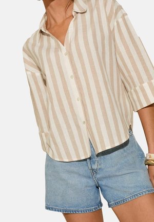 Vrouw die een beige en wit verticaal gestreept overhemd met driekwart mouwen en lichtblauwe spijkershorts draagt.